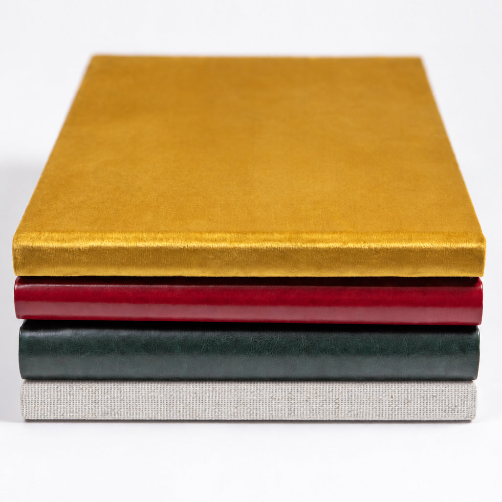 Stack of handcrafted wedding albums in dijon yellow velvet, red vintage and hunter green leatherette, and Cappuccino linen covers